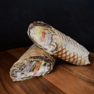 Kebab w tortilli na drewnianej desce, przekrojony na pół, prezentujący soczyste mięso, świeże warzywa (sałata, pomidor, ogórek, czerwona cebula) i kremowy sos. Idealny Kebab Kraków dla każdego smakosza, szukającego najlepszych wrażeń kulinarnych w Krakowie